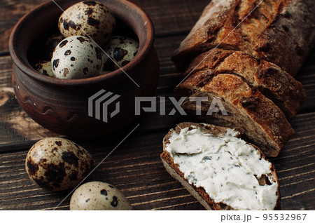 dark buckwheat bread is spread with cottage cheese with herbs in a cut on a wooden table near quail eggs in a clay plate in a rustic style. Snack and breakfast concept. dark buckwheat bread is spread with cottage cheese with herbs in a cut on a wooden table near quail eggs in a clay plate in a rustic style. Snack and breakfast concept. 95532967