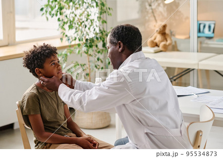 African pediatrician examining sore throat of little patient during medical exam at office African pediatrician examining sore throat of little patient during medical exam at office 95534873
