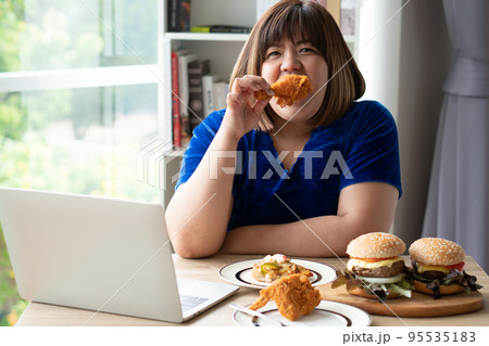 Hungry overweight woman holding Fried Chicken, hamburger on a wooden plate and Pizza on table, During work from home, gain weight problem. Concept of binge eating disorder (BED). Hungry overweight woman holding Fried Chicken, hamburger on a wooden plate and Pizza on table, During work from home, gain weight problem. Concept of binge eating disorder (BED). 95535183