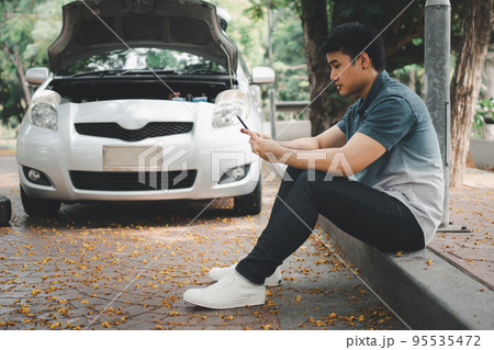 Asian man using smartphone for assistance after a car breakdown on street. Concept of vehicle engine problem or accident and emergency help from Professional mechanic 95535472