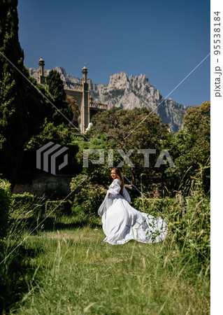 a beautiful woman with long brown hair and long white dress stay along a path along beautiful bushes in the park rear view a beautiful woman with long brown hair and long white dress stay along a path along beautiful bushes in the park rear view 95538184