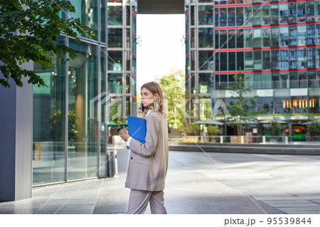 Corporate woman in business suit, walking in city center with work papers and mobile phone, calling someone 95539844