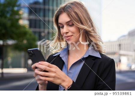 Close up portrait of young corporate woman in black suit, holds smartphone, texting message while standing on street, using mobile application Close up portrait of young corporate woman in black suit, holds smartphone, texting message while standing on street, using mobile application 95539861