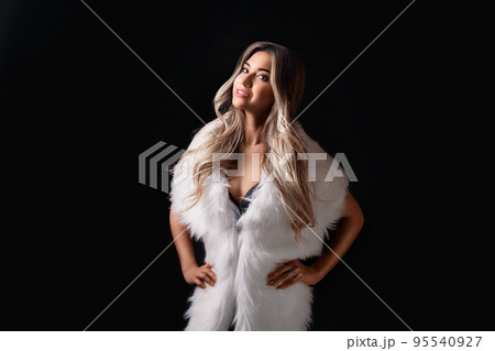 Pretty blonde girl posing in luxurious white fur cape while standing on black background. Fashion 95540927