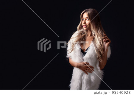 Beautiful blonde girl posing in luxurious white fur cape while standing on black background. Fashion 95540928