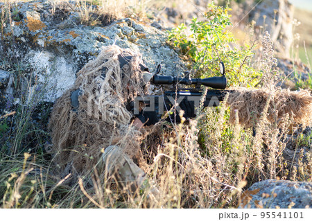 sniper in ghillie (camouflage) suit with a high-precision rifle with optic scope in a combat position in the mountains 95541101