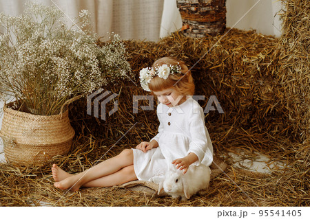 A cute and beautiful little girl is sitting in the hayloft with a rabbit A cute and beautiful little girl is sitting in the hayloft with a rabbit 95541405