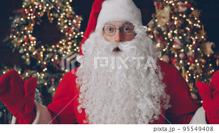 Portrait of man in traditional Santa Claus costume and glasses looking at camera near Christmas tree in dark illuminated and decorated room. Holidays and people concept. 95549408