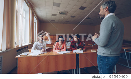 Small group of students is talking to male teacher, young man is raising hand and asking question, smiling educator is listening and gesturing. Learning and teaching concept. Small group of students is talking to male teacher, young man is raising hand and asking question, smiling educator is listening and gesturing. Learning and teaching concept. 95549409