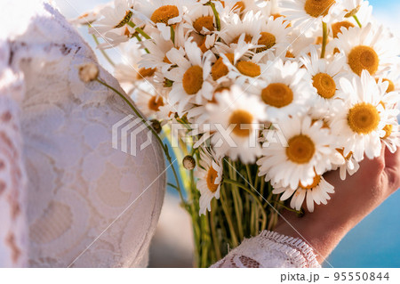 summer bouquet of field daisies in the hands of a bride in white dress. warm sunset time on the background of the sea. Copy space. The concept of calmness, silence and unity with nature. summer bouquet of field daisies in the hands of a bride in white dress. warm sunset time on the background of the sea. Copy space. The concept of calmness, silence and unity with nature. 95550844