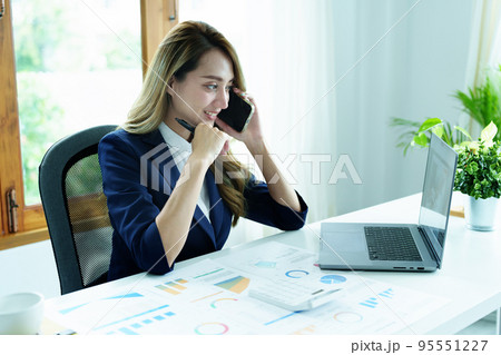 Portrait of a young Asian woman showing a smiling face as she uses her phone, computer and financial documents on her desk in the early morning hours. Portrait of a young Asian woman showing a smiling face as she uses her phone, computer and financial documents on her desk in the early morning hours. 95551227