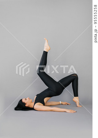 Young woman fitness trainer or yoga performs an exercise lying on her shoulder blades and lifting one leg at the top on a gray background 95555258