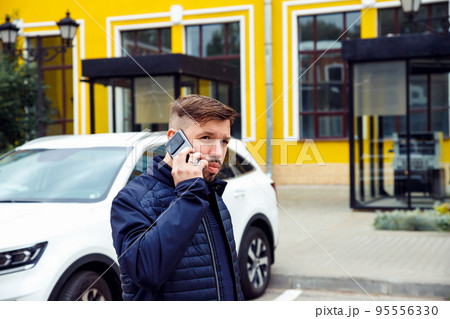 Young Caucasian businessman 34 years old man with goatee beard talks on smartphone on street near white car. Waist up lifestyle business portrait, success concept. 95556330