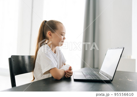 Side view of cute blonde little girl with broken hand wrapped in white plaster bandage sitting at table with laptop computer looking to screen in light room. Concept of child insurance and healthcare. Side view of cute blonde little girl with broken hand wrapped in white plaster bandage sitting at table with laptop computer looking to screen in light room. Concept of child insurance and healthcare. 95556610