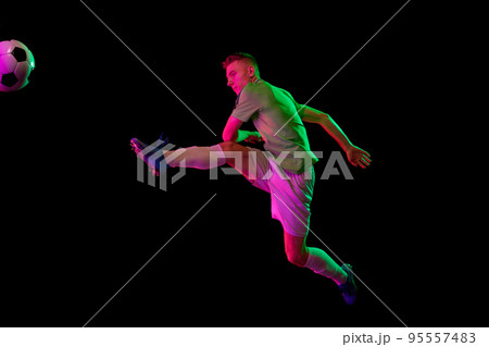 Sport in action. One man professional soccer player training with football ball isolated on dark background in neon light filter. Sport, speed, power and energy 95557483