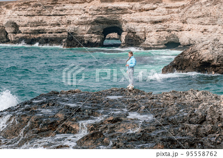 A woman in a blue jacket stands on a rock above a cliff above the sea and looks at the raging ocean. Girl traveler rests, thinks, dreams, enjoys nature. Peace and calm landscape, windy weather. A woman in a blue jacket stands on a rock above a cliff above the sea and looks at the raging ocean. Girl traveler rests, thinks, dreams, enjoys nature. Peace and calm landscape, windy weather. 95558762