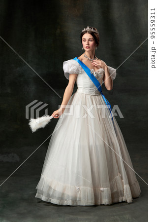 Young queen. Portrait of adorable girl in image of medieval royal person in renaissance style dress isolated on dark background. Comparison of eras, beauty, history, art 95559311