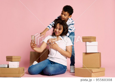 Portrait of woman with her son doing online order on phone, sitting around boxes isolated over pink studio background 95560308