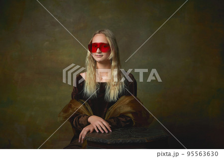 Vintage style portrait of young charming girl in medieval dress and modern red eyewear isolated on dark green background. Art, beauty, style, imitation 95563630