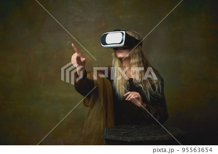 Beautiful young girl with long blonde hair wearing medieval dress and VR headset over dark vintage background. Retro style, art, fashion, comparison of eras concept. 95563654