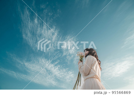 青空のもとで花を掲げる白いワンピースの女性 95566869