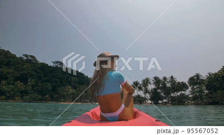 Sexy young woman in blue swimsuit and hat swings feet in water on pink canoe on sea against hills backside view. Sexy young woman in blue swimsuit and hat swings feet in water on pink canoe on sea against hills backside view. 95567052