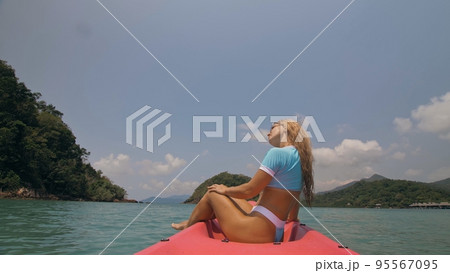 Pretty young blonde woman in stylish swimsuit and sunglasses sits on pink canoe sailing along sea against hills. 95567095