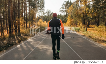 Training an athlete on the roller skaters. Biathlon ride on the roller skis with ski poles, in the helmet. Autumn workout. Roller sport. The athlete goes and holds sports equipment in his hand. 95567554