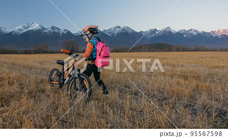 One caucasian children walk with bike in wheat field. Little girl walking black orange cycle on background of beautiful snowy mountains. Biker stand with backpack and helmet. One caucasian children walk with bike in wheat field. Little girl walking black orange cycle on background of beautiful snowy mountains. Biker stand with backpack and helmet. 95567598