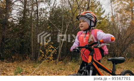 One caucasian children walk with bike in autumn park. Little girl walking black orange cycle in forest. Kid goes do bicycle sports. Biker motion ride with backpack and helmet. Mountain bike hard tail. One caucasian children walk with bike in autumn park. Little girl walking black orange cycle in forest. Kid goes do bicycle sports. Biker motion ride with backpack and helmet. Mountain bike hard tail. 95567701