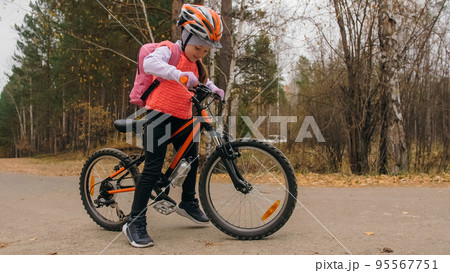 One caucasian children walk with bike in autumn park. Little girl walking black orange cycle in forest. Kid goes do bicycle sports. Biker motion ride with backpack and helmet. Mountain bike hard tail. 95567751