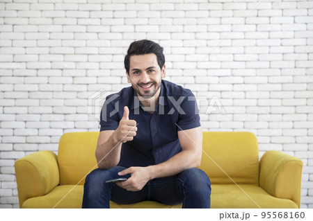 Lifestyle portrait of attractive handsome man with pleasant smile relaxing and sitting on yellow sofa at living room 95568160