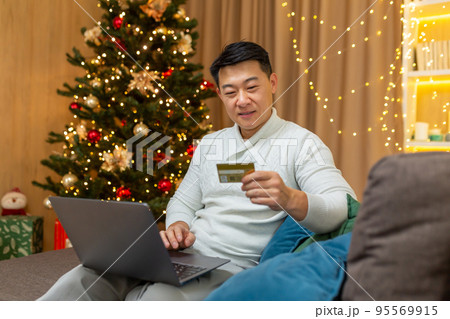 Successful businessman on christmas at home sitting on sofa in living room, using laptop online shopping, asian celebrating new year near tree, choosing gifts in online store shopping credit card. 95569915