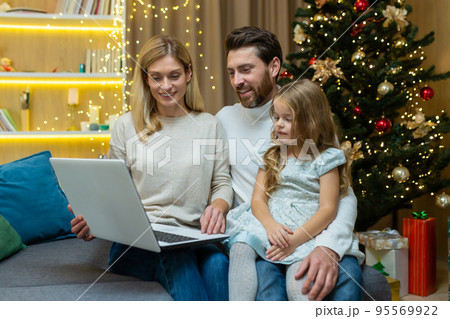 Happy family man wife and daughter celebrating new year and Christmas together, couple sitting on sofa at home near Christmas tree with laptop, watching video online together. 95569922