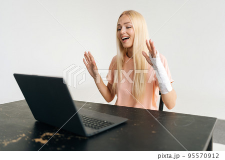 Portrait of positive injured blonde young woman with broken arm wrapped in plaster bandage having video call or online consultation using laptop computer, on white isolated background. Portrait of positive injured blonde young woman with broken arm wrapped in plaster bandage having video call or online consultation using laptop computer, on white isolated background. 95570912
