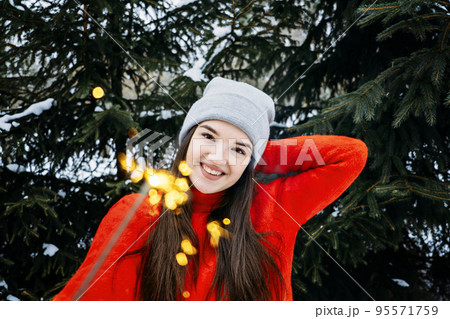 Outdoor portrait of happy cute Latina girl in winter hat and red sweater posing with sparkler against Christmas trees with snow background. Brunette woman having fun in winter holidays. 95571759