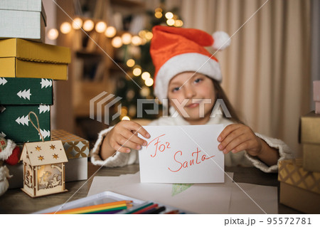 Little preschool child girl in red hat with letter to Santa Claus sitting at table with gifts 95572781