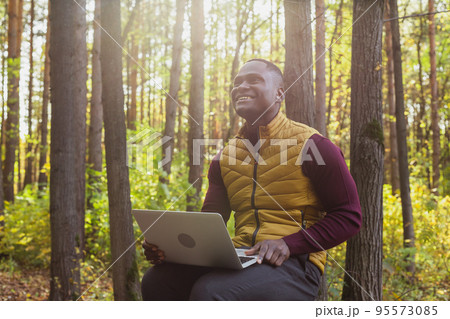 African american man sitting in city park on a bench with laptop studying online outdoors. Man freelancer learn working remotely in street e-learning 95573085