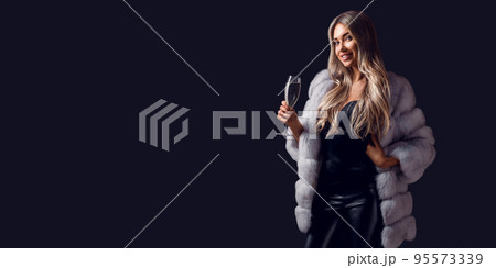 Gorgeous young woman in fur coat holds champagne in hand stands while smiling celebrating something 95573339