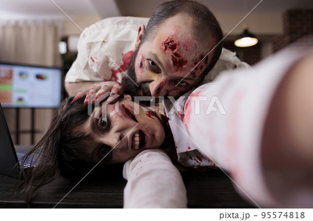 Scary horror zombies biting wounds in office, looking creepy cruel and terrifying at desk. Walking dead evil couple of corpses with blood on scars, spooky dramatic eerie nightmare with monsters. 95574818