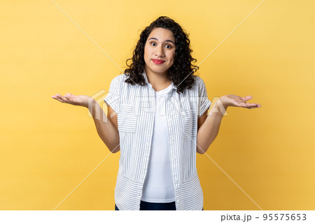 I am not sure. Portrait of ambiguous woman with dark wavy hair standing with raised arms, looking away and don't know what to do. Indoor studio shot isolated on yellow background. 95575653