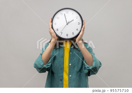 Portrait of unknown woman holding wall clock hiding her face, time management, schedule and meeting appointment, wearing casual style jacket. Indoor studio shot isolated on gray background. Portrait of unknown woman holding wall clock hiding her face, time management, schedule and meeting appointment, wearing casual style jacket. Indoor studio shot isolated on gray background. 95575710