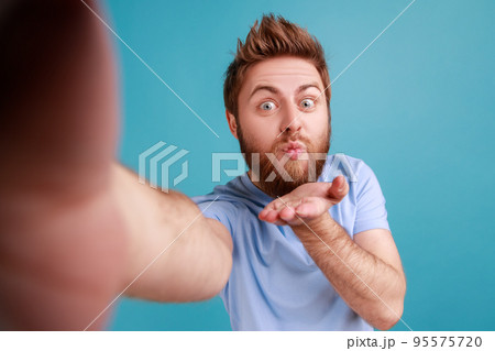 Portrait of romantic flirting bearded handsome man blogger making selfie POV, sending air kisses to followers while having livestream. Indoor studio shot isolated on blue background. 95575720