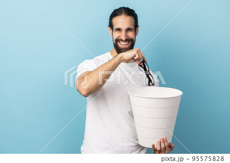 Portrait of man with beard wearing white T-shirt throwing out eyeglasses into rubbish bin, treat his eyesight, looking smiling at camera. Indoor studio shot isolated on blue background. 95575828