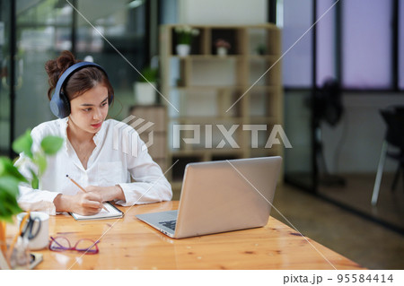 Portrait of a beautiful woman using a computer, earphone and notebook during a video conference 95584414