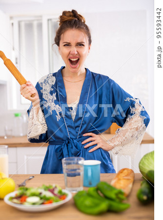 Angry woman screaming and waving rolling pin in home kitchen 95593442