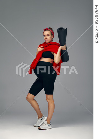 Pregnant woman holding yoga mat in hands, going for sport while awaiting baby, looking smiling at camera and touching her belly, standing against gray wall. 95597644