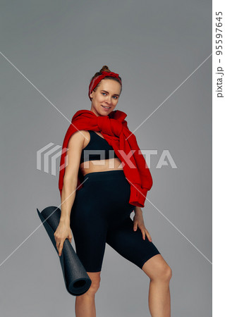 Pregnant woman holding yoga mat in hands, going for sport while awaiting baby, looking smiling at camera and touching her belly, standing against gray wall. Pregnant woman holding yoga mat in hands, going for sport while awaiting baby, looking smiling at camera and touching her belly, standing against gray wall. 95597645