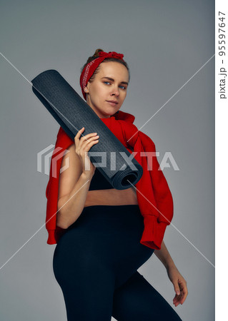 Pregnant woman holding yoga mat in hands, going for sport while awaiting baby, looking smiling at camera and touching her belly, standing against gray wall. Pregnant woman holding yoga mat in hands, going for sport while awaiting baby, looking smiling at camera and touching her belly, standing against gray wall. 95597647