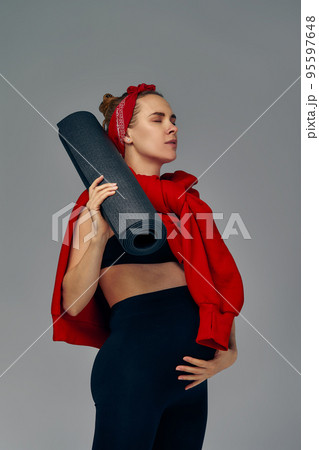 Pregnant woman holding yoga mat in hands, going for sport while awaiting baby, looking smiling at camera and touching her belly, standing against gray wall. Pregnant woman holding yoga mat in hands, going for sport while awaiting baby, looking smiling at camera and touching her belly, standing against gray wall. 95597648